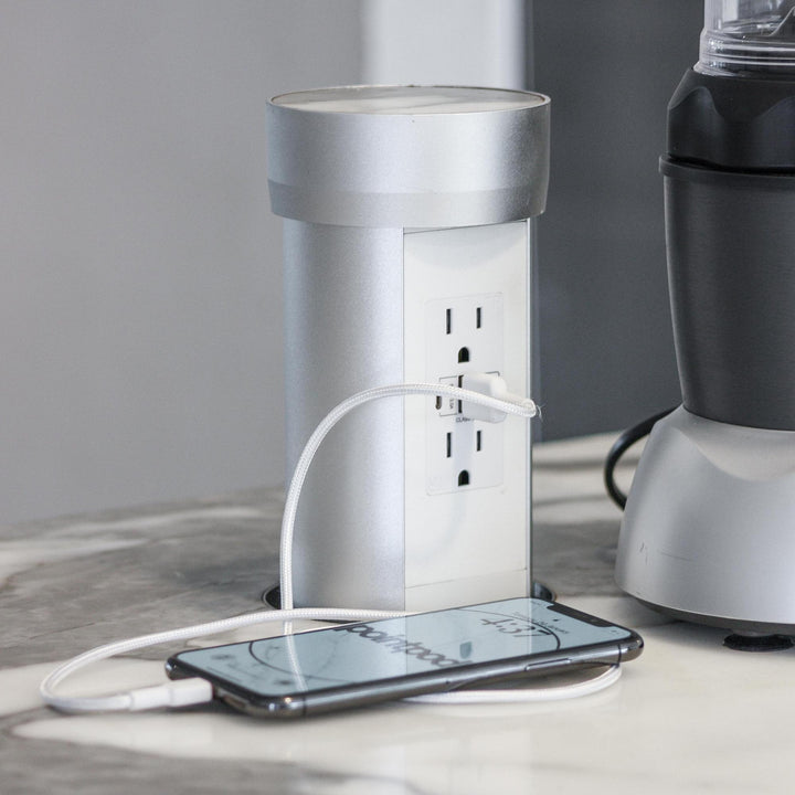 White point pod power outlet installed in a countertop, featuring two AC outlets and two USB ports. A smartphone is connected and charging, showing its versatility in powering multiple devices.